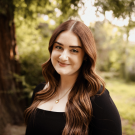 A photo of a woman with brown hair, a tree in the background.